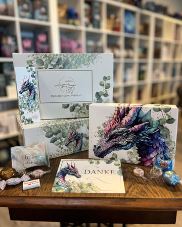 Das Bild zeigt auf einer alten Nähmaschine und vor den Bücherregalen im Drachennest eine Buchbox von oben, eine Buchbox von der Seite, ein in ein Drachen-Poster verpacktes Buch und eine Postkarte mit einem Drachen und dem Schriftzug "Danke" sowie ein paar Süßigkeiten. Die Hintergründe von Box, Poster und Karte sind weiß, der je darauf gezeigte Drache ist in Schattierungen der Farben Violett, Lila und Grün gestaltet. Schuppen und Eukalyptus-Blätter ziehen sich zusätzlich über Box, Poster und Karte.